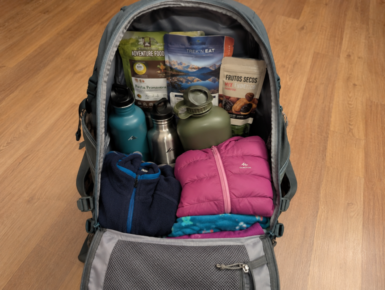 Mochila de día de 30 litros abierta mostrando comida de camping, cantimploras y ropa de abrigo infantil